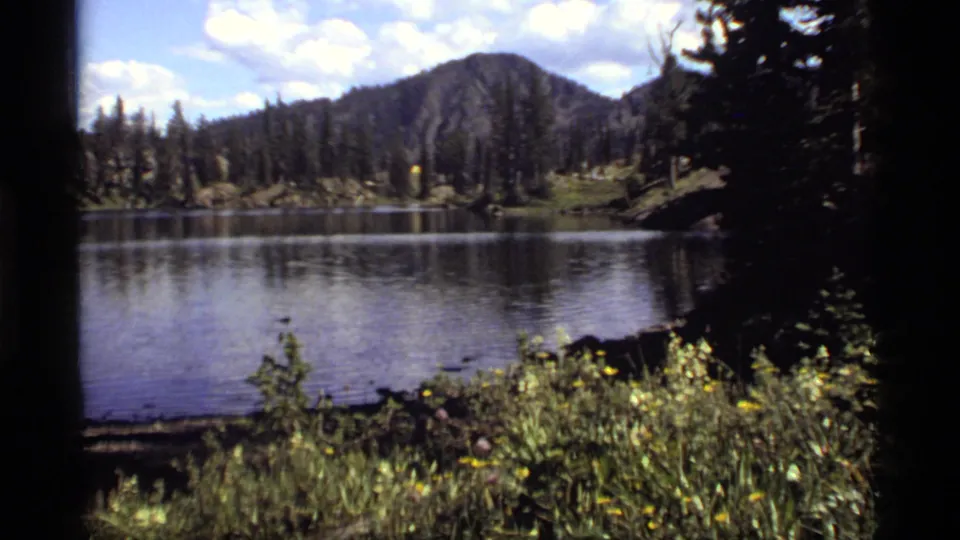 A handheld 8mm lake view shows Sapphire Lake with dark pines, distant mountain, and sunlit foreground vegetation.