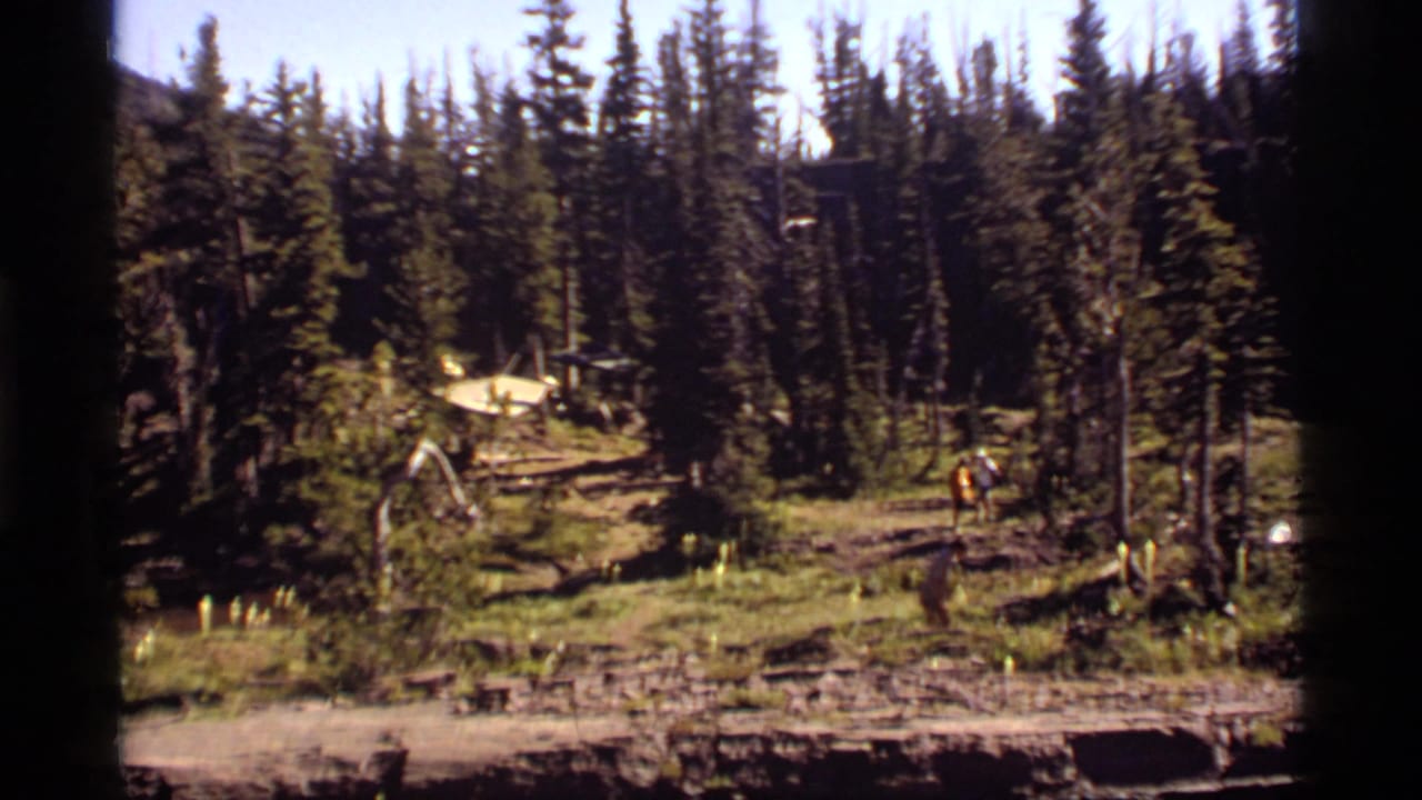 1977 Sapphire Lake, Montana frame with pine trees, a dirt trail, a yellow tent, and a distant visitor in the forest clearing.