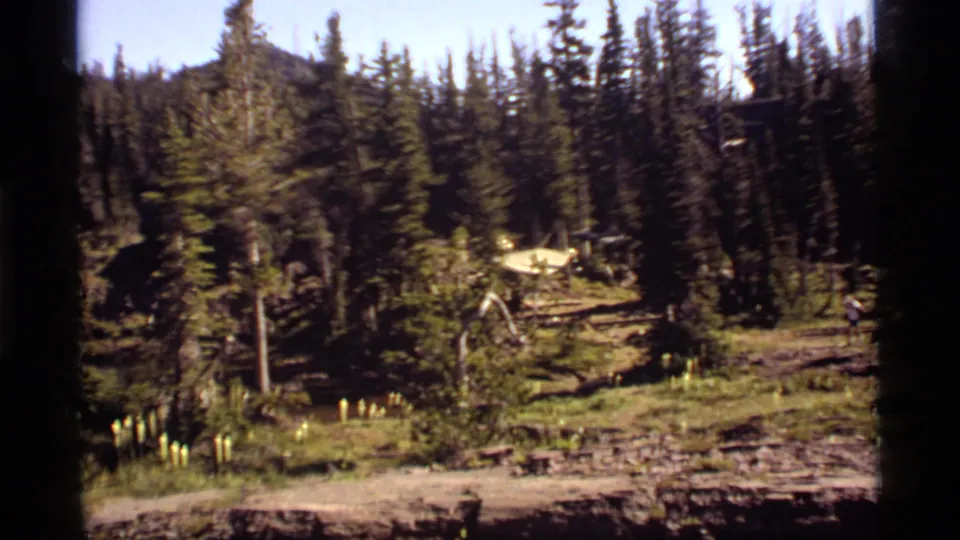 1977 Sapphire Lake, Montana frame showing a dirt path, tall pines, a yellow tent, and distant visitors at the edge of the clearing.