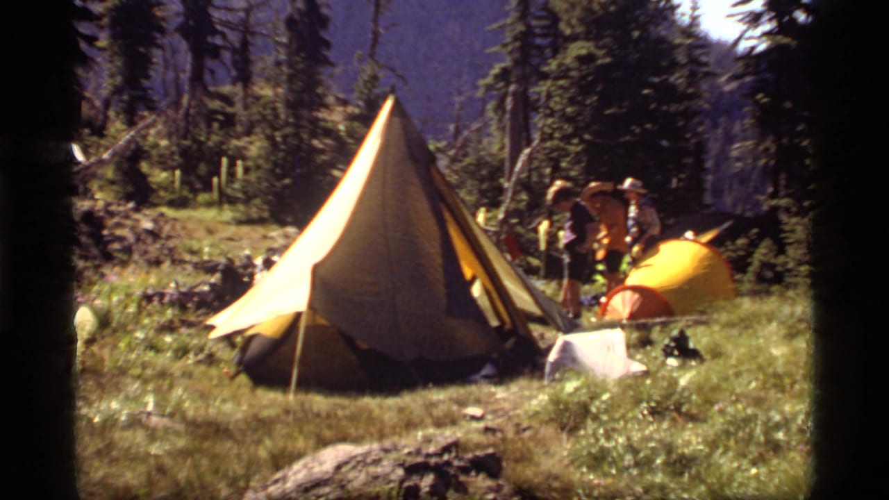 1977 Sapphire Lake campsite frame with a large yellow A-frame tent, a smaller yellow dome tent, campers in hats, meadow grass, and surrounding evergreens.