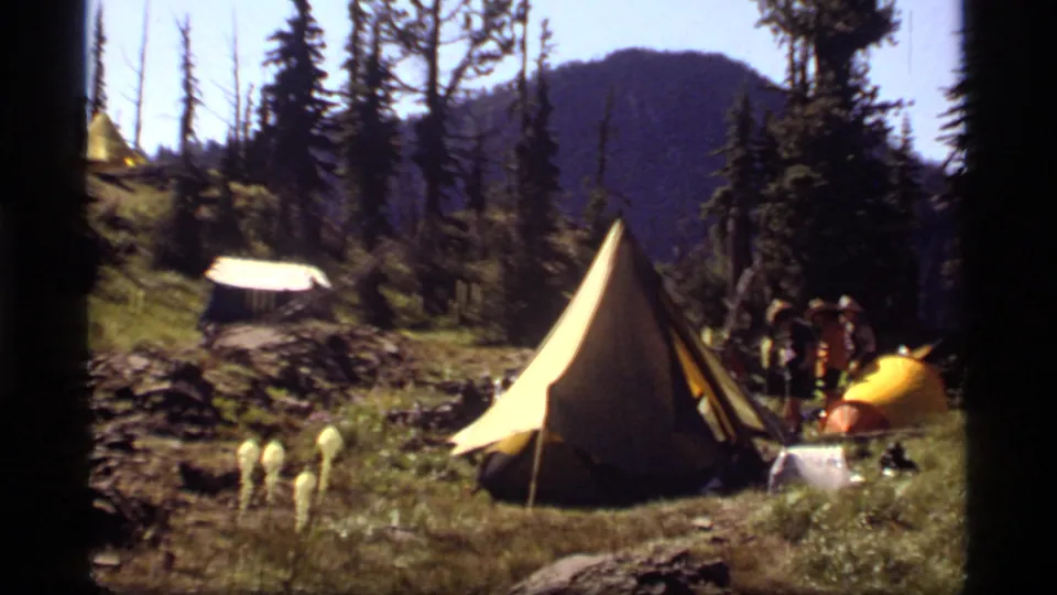 Sapphire Lake, Montana campsite view with a yellow A-frame tent, campers beside a small tent, a white tarp or table cover, wildflowers, trees, and mountains.