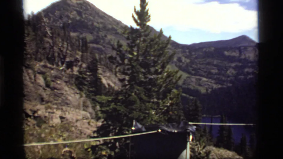 1977 8mm mountain view near Sapphire Lake with a rope line, dark shelter edge, pine tree, rocky slope, and water in the distance.