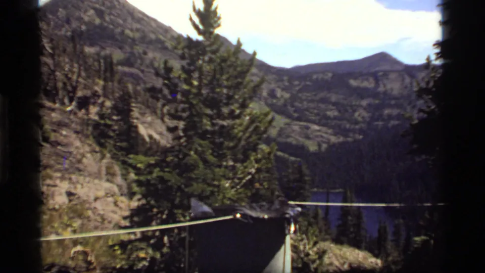 Sapphire Lake, Montana mountain view showing a rope line, dark shelter edge, pine tree, rocky terrain, forested slopes, and distant water.
