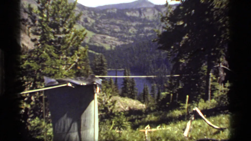 Handheld 1977 8mm campsite view with a dark shelter in the foreground and Sapphire Lake partly visible through trees.