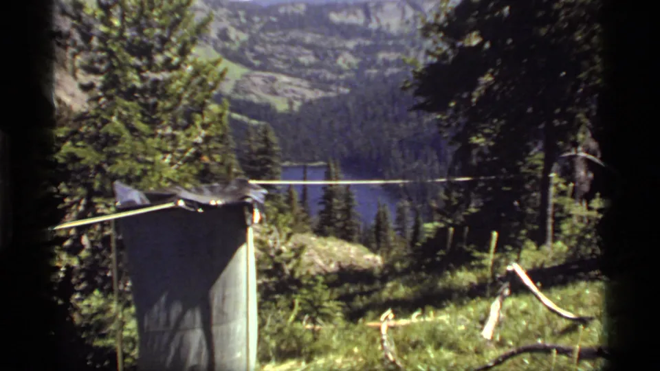 1977 8mm view near Sapphire Lake, Montana, with evergreen trees, a camp structure, distant water, and mountain slopes.