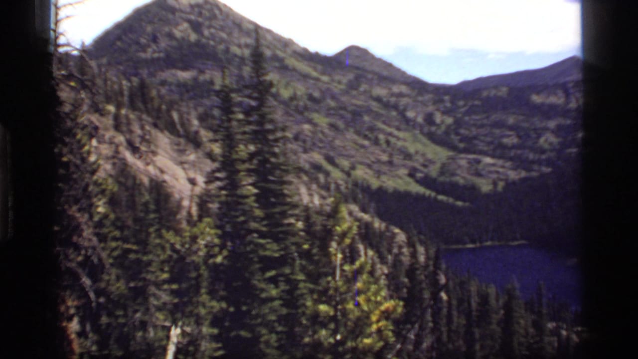 1977 8mm Montana mountain view showing Sapphire Lake below evergreen forest, rocky slopes, and rounded peaks.