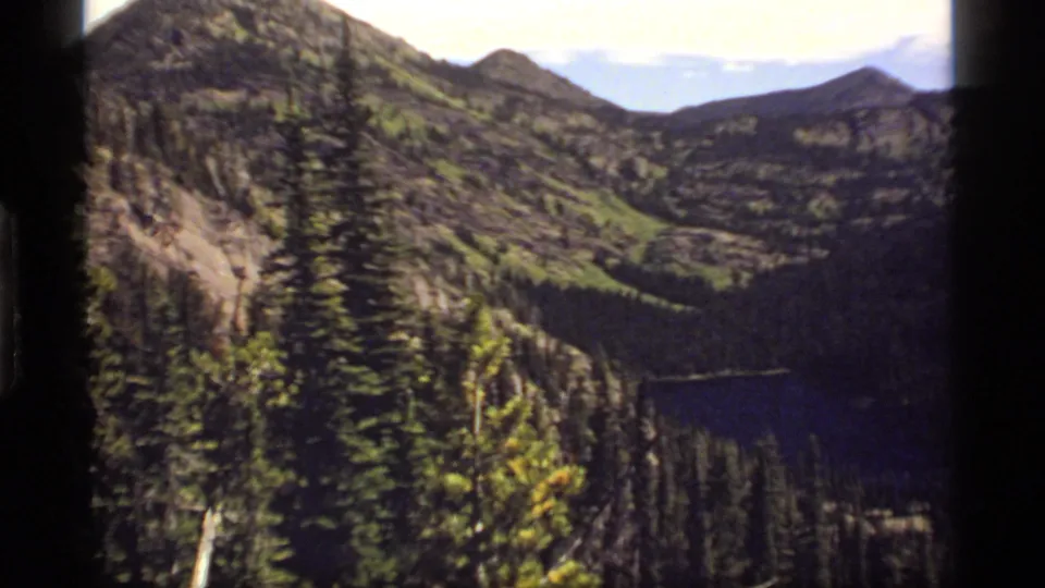1977 8mm frame of Sapphire Lake, Montana, with dense conifers, blue water, rugged slopes, and soft home-movie color.