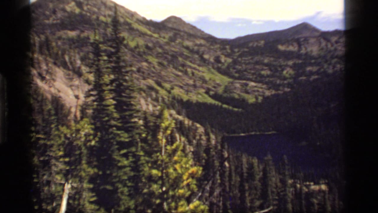 1977 8mm view over Sapphire Lake, Montana, with evergreen forest, rocky slopes, distant peaks, and dark film-frame edges.