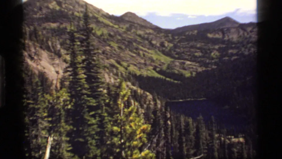 1977 8mm view over Sapphire Lake, Montana, with evergreen forest, rocky slopes, distant peaks, and dark film-frame edges.