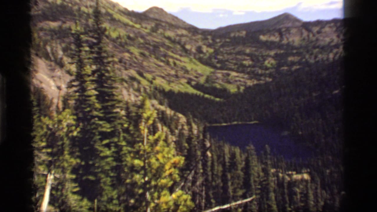 Mountain valley with evergreen trees, rocky slopes, and a lake partly hidden below.