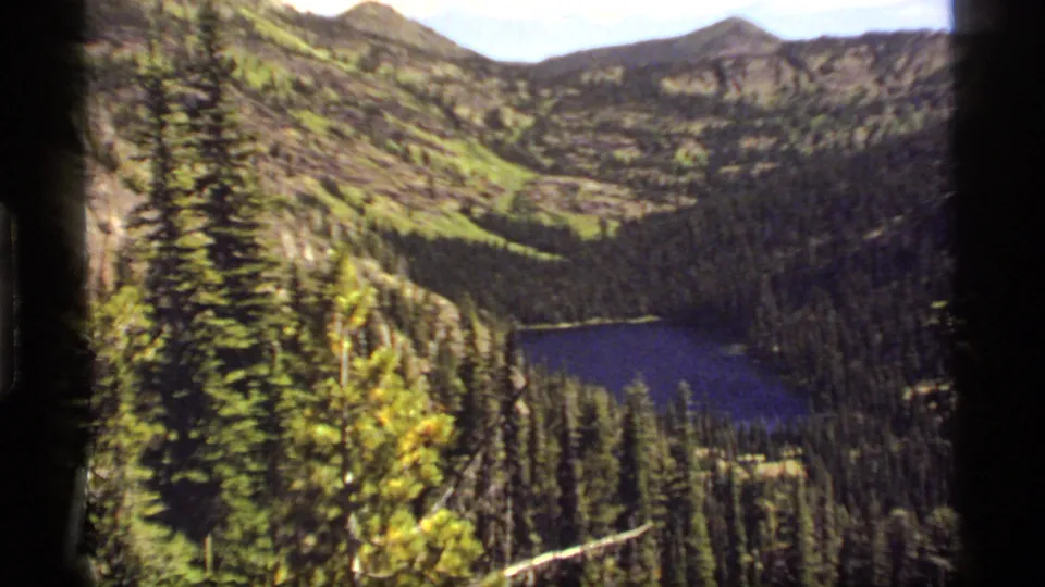 1977 Montana home-movie view of Sapphire Lake below dense evergreens and rocky green-brown mountain terrain.