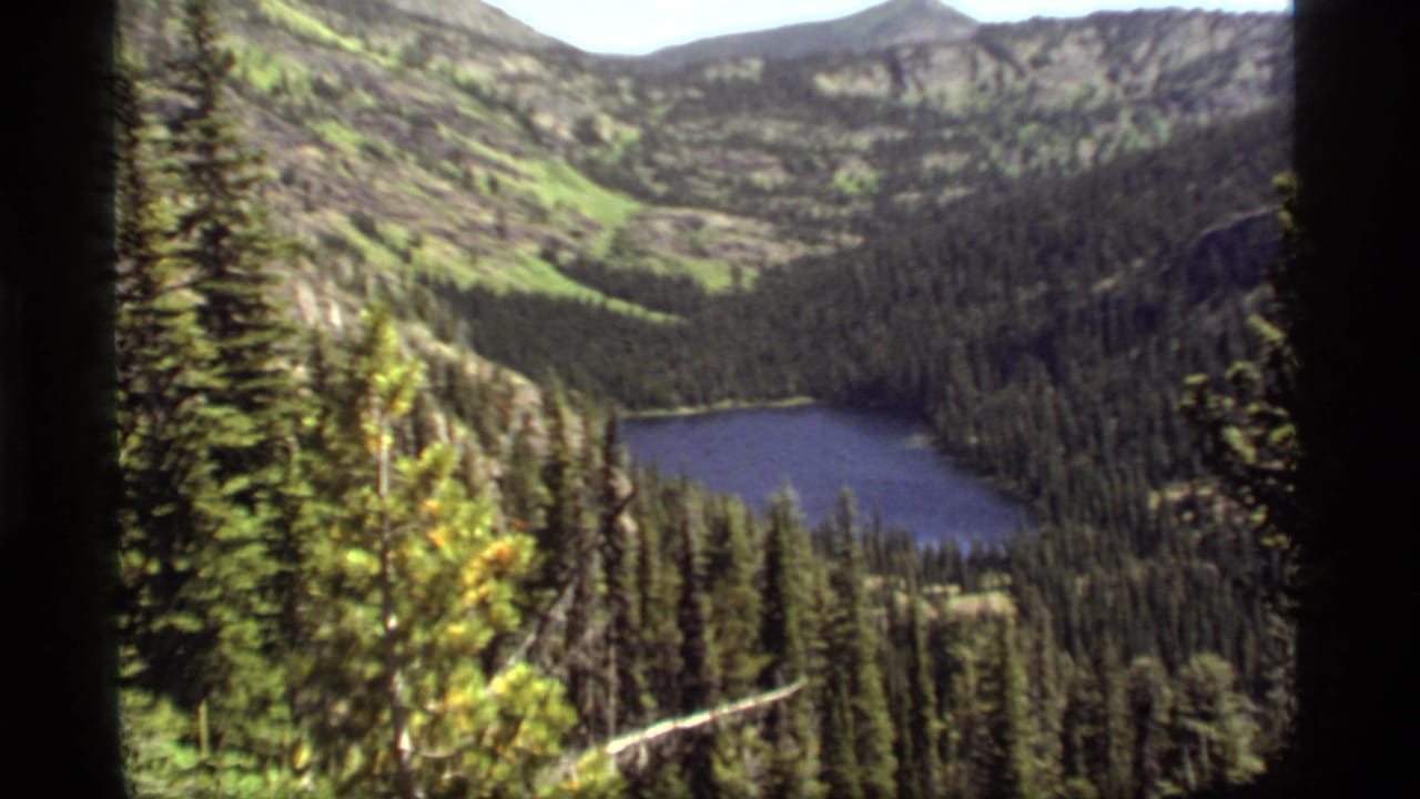 1977 8mm view of Sapphire Lake in Montana, seen from above through evergreen forest and mountain slopes.