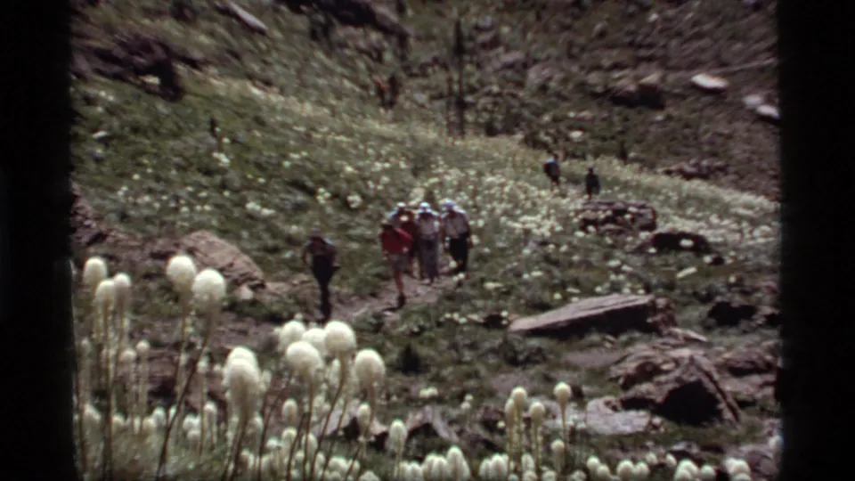 A wider 1977 home-movie view shows hikers crossing a Sapphire Lake meadow with bear grass, rocks, and forested terrain.