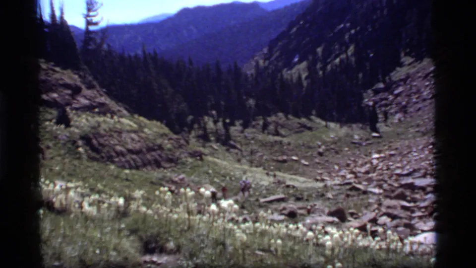 Handheld 1977 8mm frame of hikers moving through the Sapphire Lake mountain valley, with wildflowers and rocky ground visible.