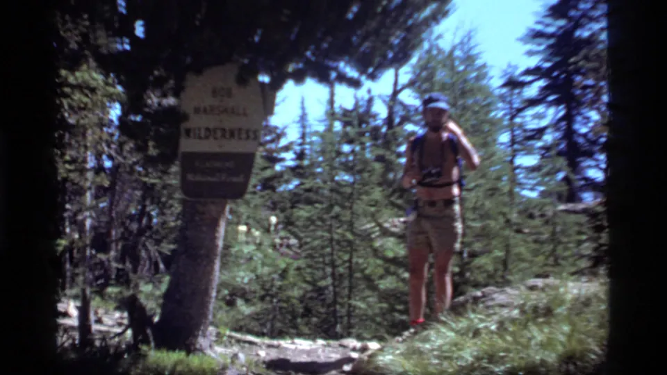 A shirtless adult stands with binoculars near a Bob Marshall Wilderness sign along a forest path in Montana, 1977.