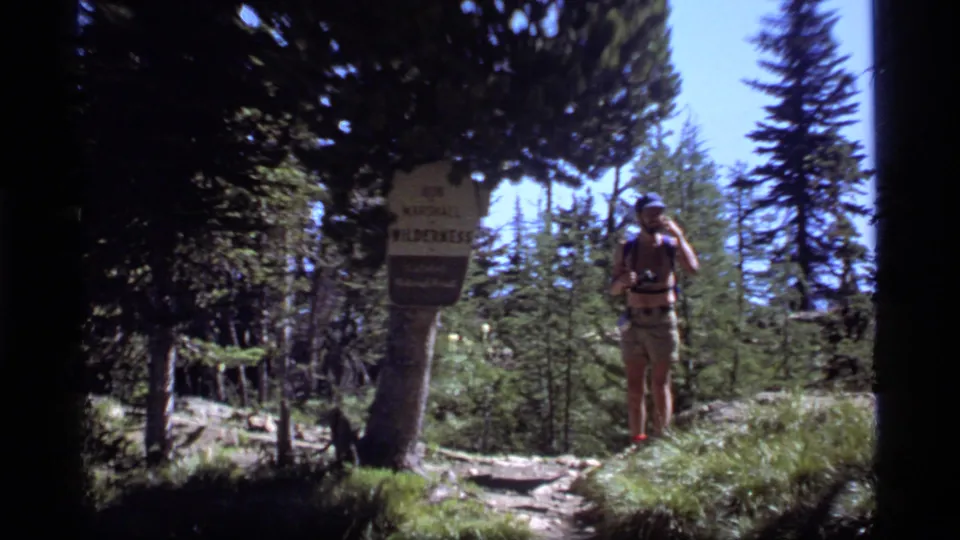 A visitor holding binoculars stands beside a Bob Marshall Wilderness sign on a sunlit Montana forest path in 1977 8mm footage.