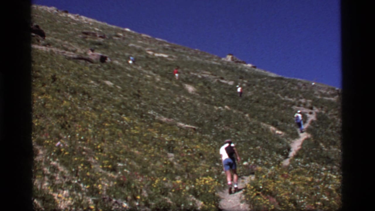 Hikers in casual summer clothing ascend a sunny Sapphire Lake, Montana hillside path in 1977 8mm footage.