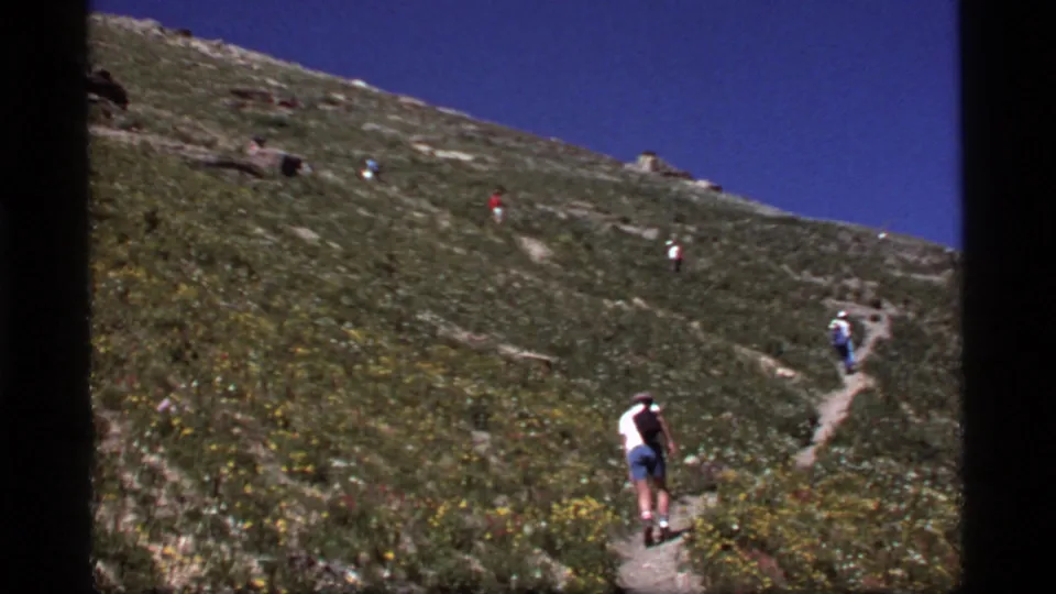 Hikers in casual summer clothing ascend a sunny Sapphire Lake, Montana hillside path in 1977 8mm footage.
