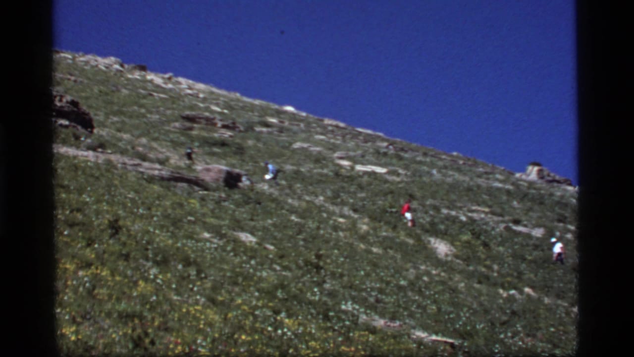 1977 8mm home-movie frame from Sapphire Lake, Montana, showing distant hikers crossing a grassy mountain slope beneath a deep blue sky.