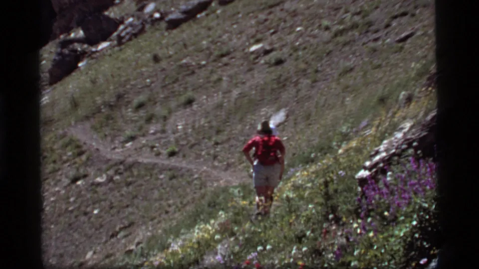 A hiker in a red shirt and backpack walks away on a winding grassy trail in the Sapphire Lake, Montana mountain setting.