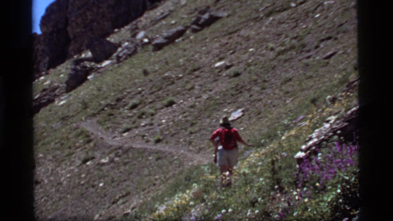 At Sapphire Lake, Montana in 1977, a hiker in red and light shorts walks away along a rocky mountain trail bordered by wildflowers.