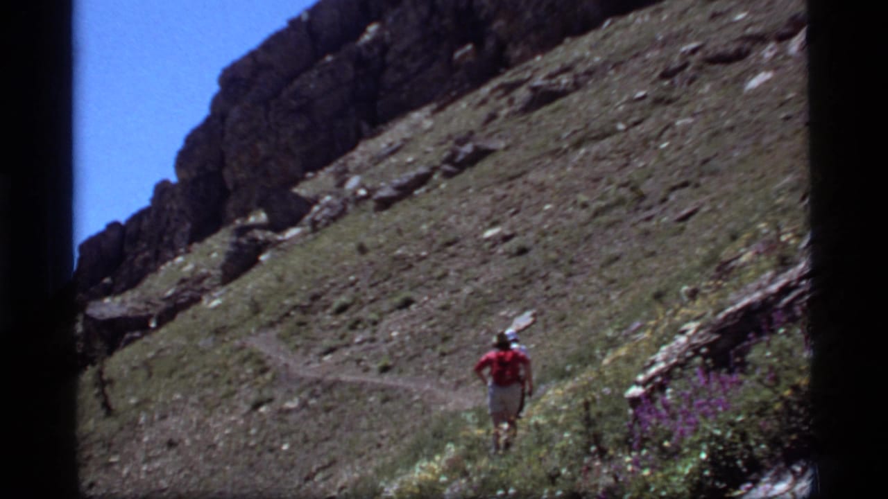 Hiker continues along a distant mountain trail beneath a rocky ridge and blue sky.