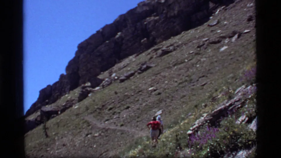 A rear-view 8mm frame shows the hiker small in the landscape, following the Sapphire Lake trail across a steep Montana slope.
