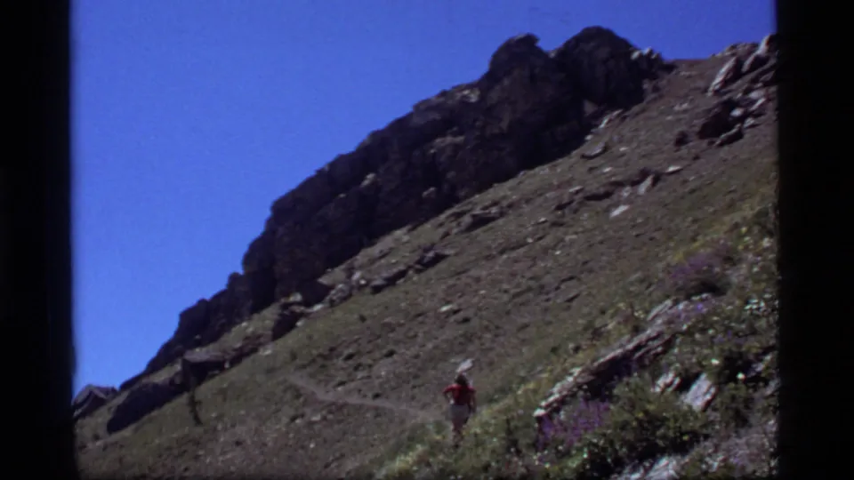 1977 8mm Montana home-movie frame showing a small hiker in red ascending a rocky trail near Sapphire Lake.