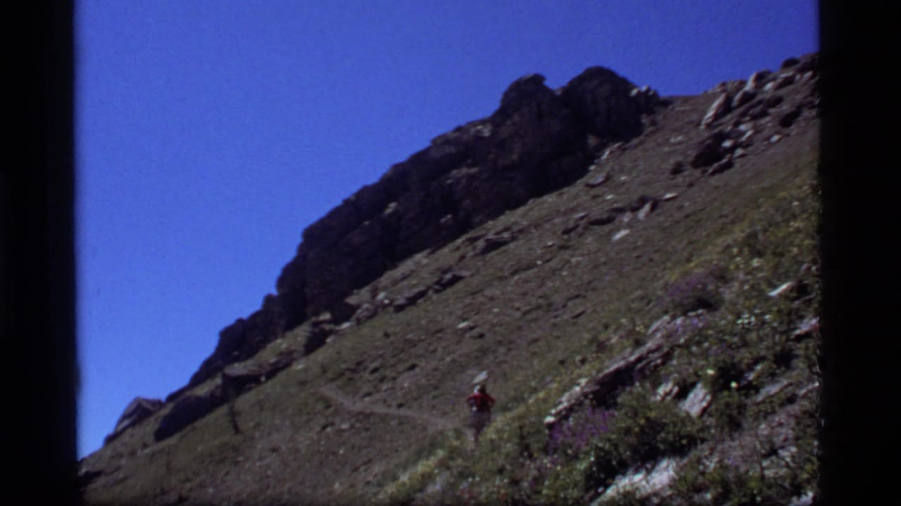 A distant hiker moves along a rugged Sapphire Lake, Montana mountain trail in 1977 8mm footage.