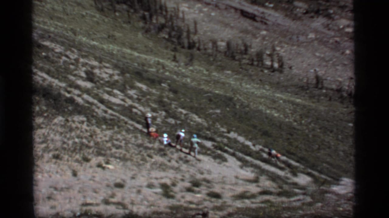 1977 8mm Montana footage showing hikers traversing a steep rocky slope near Sapphire Lake, with sparse alpine trees in the background.