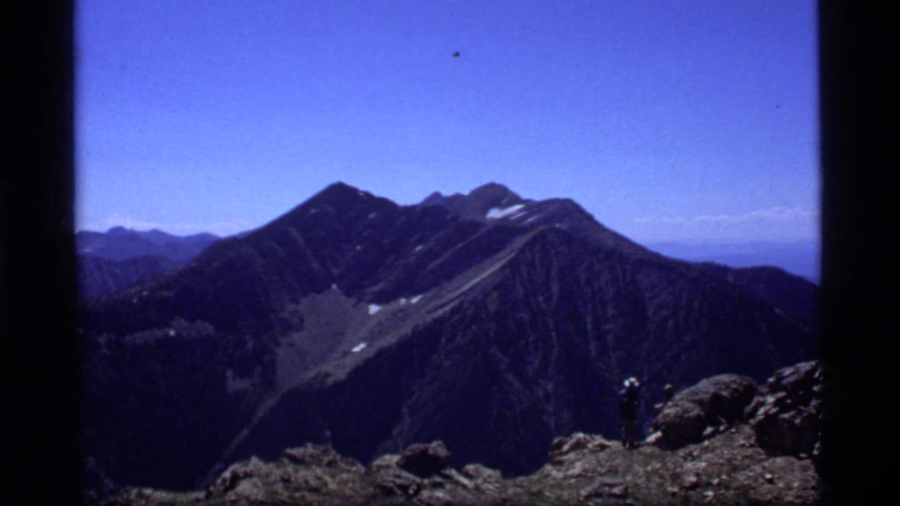 1977 8mm view near Sapphire Lake, Montana, showing rugged peaks, snow patches, blue sky, and a distant hiker on a rocky ridge.