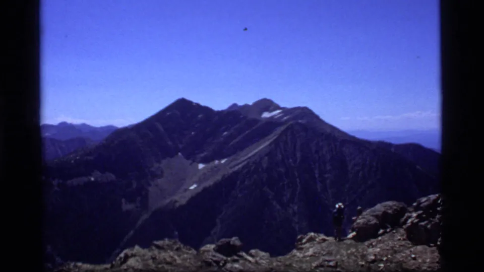 1977 8mm view near Sapphire Lake, Montana, showing rugged peaks, snow patches, blue sky, and a distant hiker on a rocky ridge.