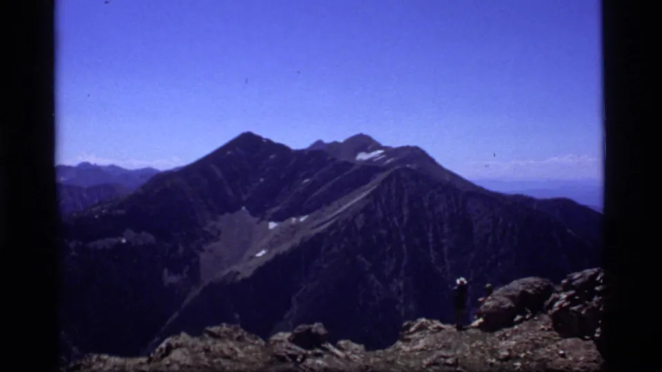 1977 8mm mountain vista near Sapphire Lake, Montana, with dark ridges, snow patches, blue sky, and a distant hiker on rocks.