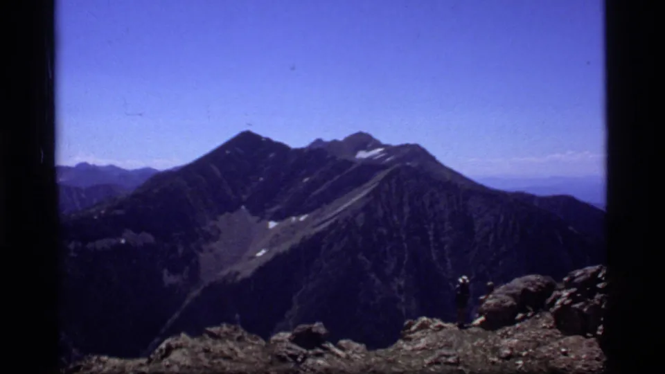 1977 8mm shot near Sapphire Lake, Montana, showing dark alpine slopes, a rocky overlook, snow patches, and a distant hiker.