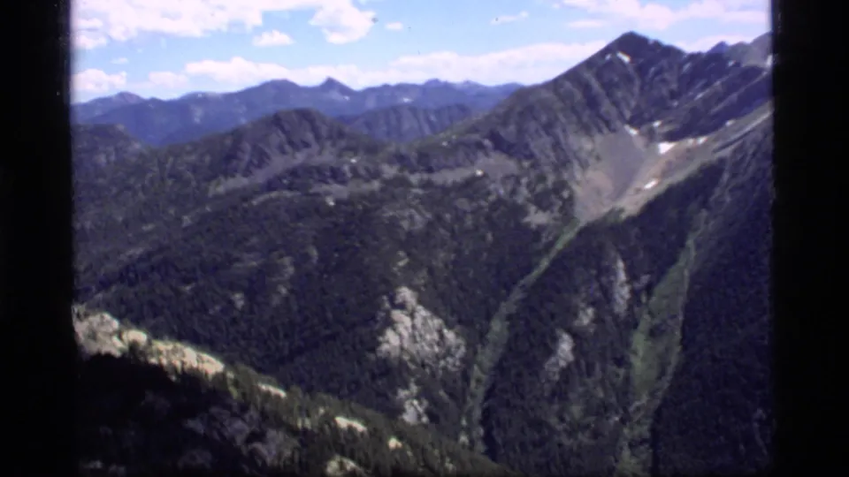1977 8mm view near Sapphire Lake, Montana, looking across a forested valley toward steep ridges with lingering snow patches.