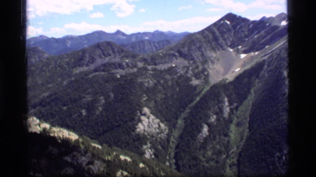 1977 8mm home-movie view of Sapphire Lake, Montana mountain ridges with forested slopes, rock faces, snow patches, and blue sky.