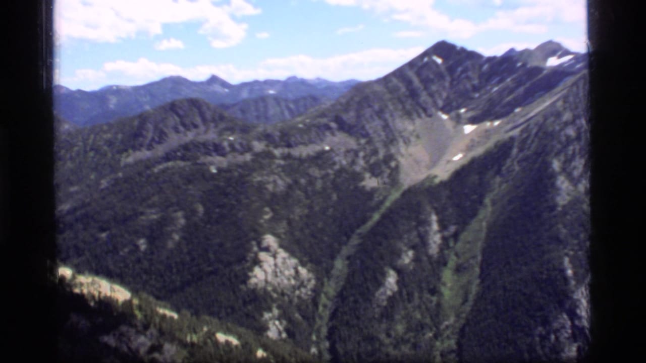 Wide alpine panorama with rugged ridgelines, forested valleys, snow traces, and dark film borders.