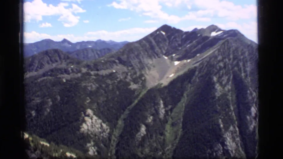 1977 Montana home-movie frame showing steep Sapphire Lake area ridges, forested slopes, rocky basin, snow remnants, and soft film grain.