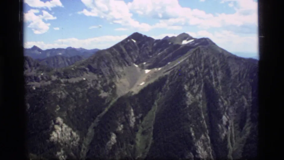 Sapphire Lake, Montana mountain peak on 8mm film, with forested slopes, exposed rock, small snow patches, and bright clouded sky.