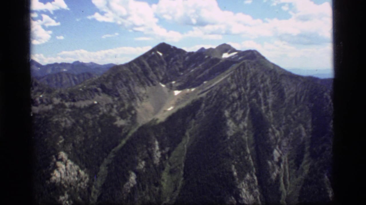 1977 8mm Montana footage showing a rugged mountain ridge, dark forested slopes, small snow patches, and a bright blue sky.