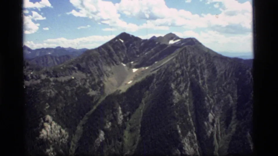 1977 8mm Montana mountain view with layered ridges, rocky slopes, forested areas, and scattered clouds above the peaks.