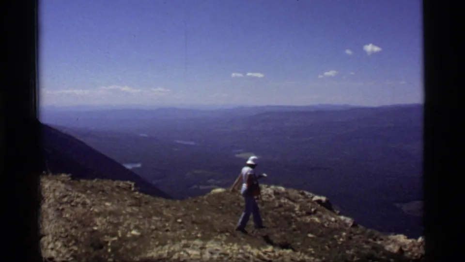 1977 8mm Montana ridge scene with a person in a white hat walking near the edge of a rocky overlook above a distant valley.