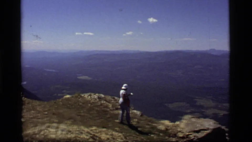 1977 8mm Montana footage showing a person in a white hat on a rocky overlook, looking across a broad valley and distant ridgelines.
