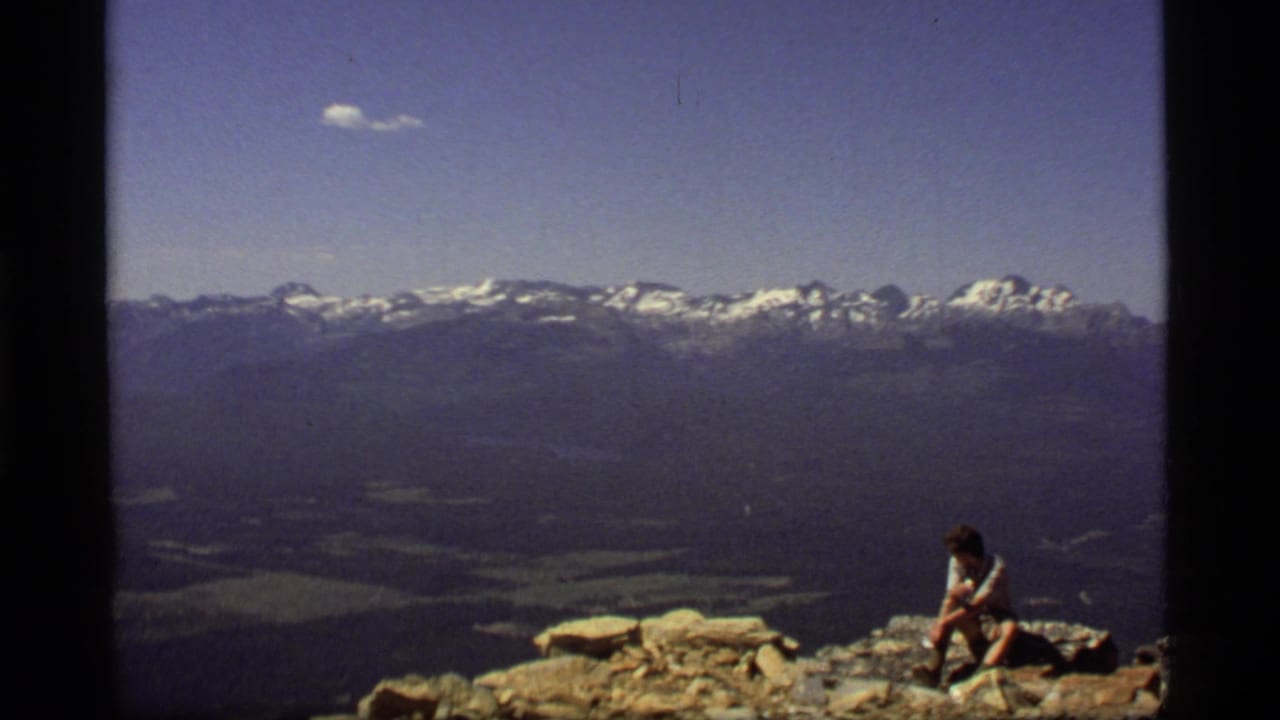 1977 8mm Montana home-movie frame showing a lone hiker resting on rocks above a broad alpine mountain view.