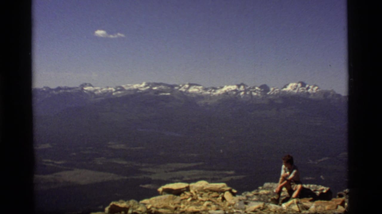 Lone hiker seated on rocks below a wide mountain range and blue sky.