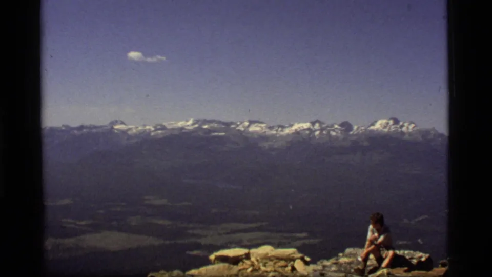 1977 8mm frame from Sapphire Lake, Montana, showing a seated hiker against a broad mountain and valley panorama.