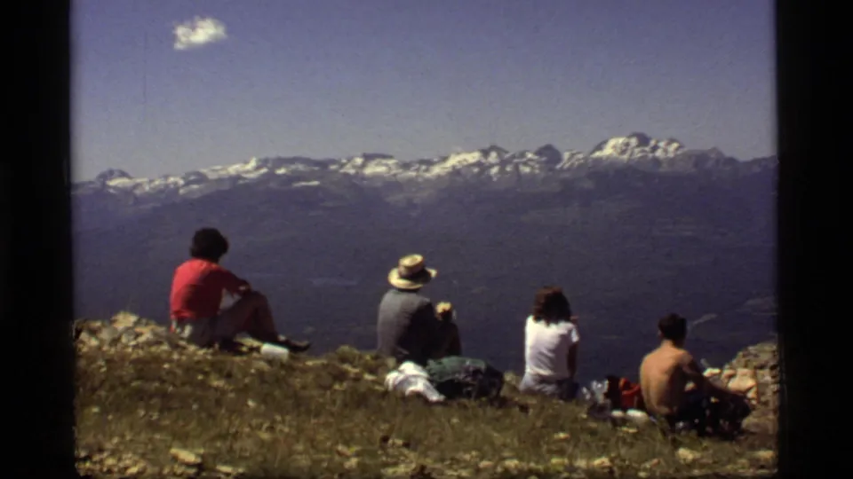 A small group sits on a Montana summit in 1977, facing a distant snow-capped mountain range under clear sky.