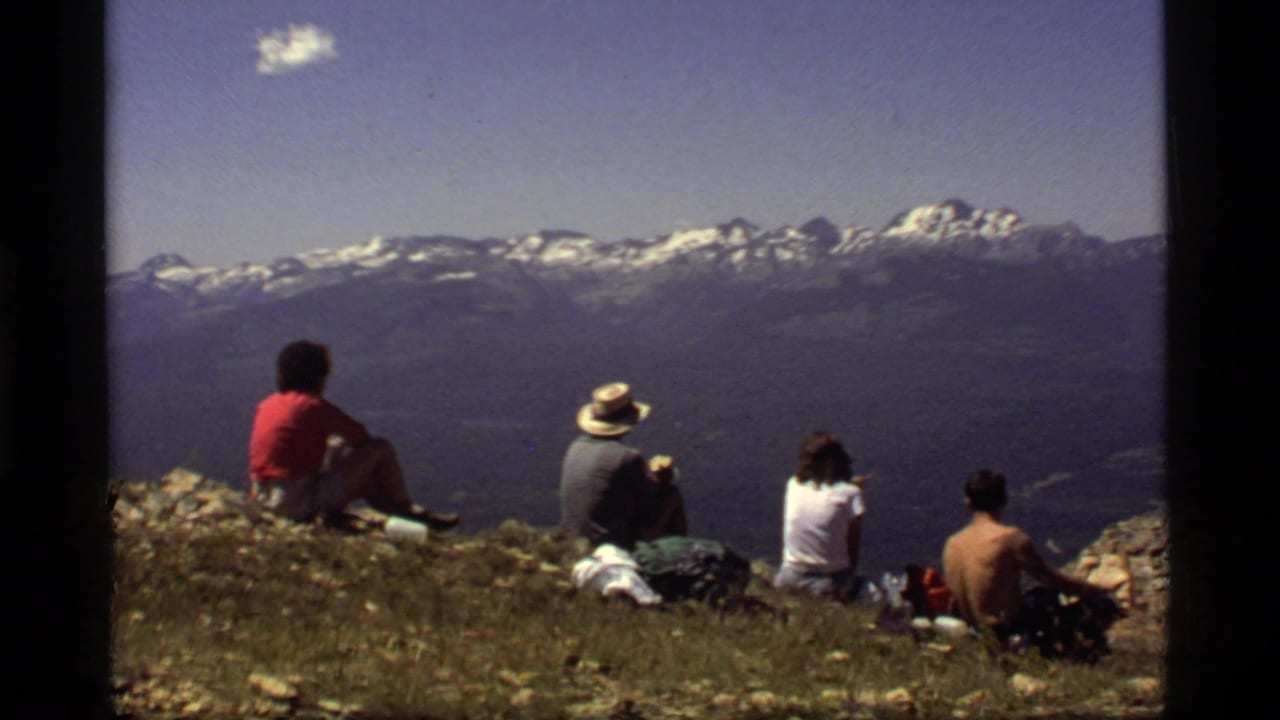 1977 8mm home-movie frame from Sapphire Lake, Montana, showing four hikers seated on a ridge and looking toward snow-capped peaks.