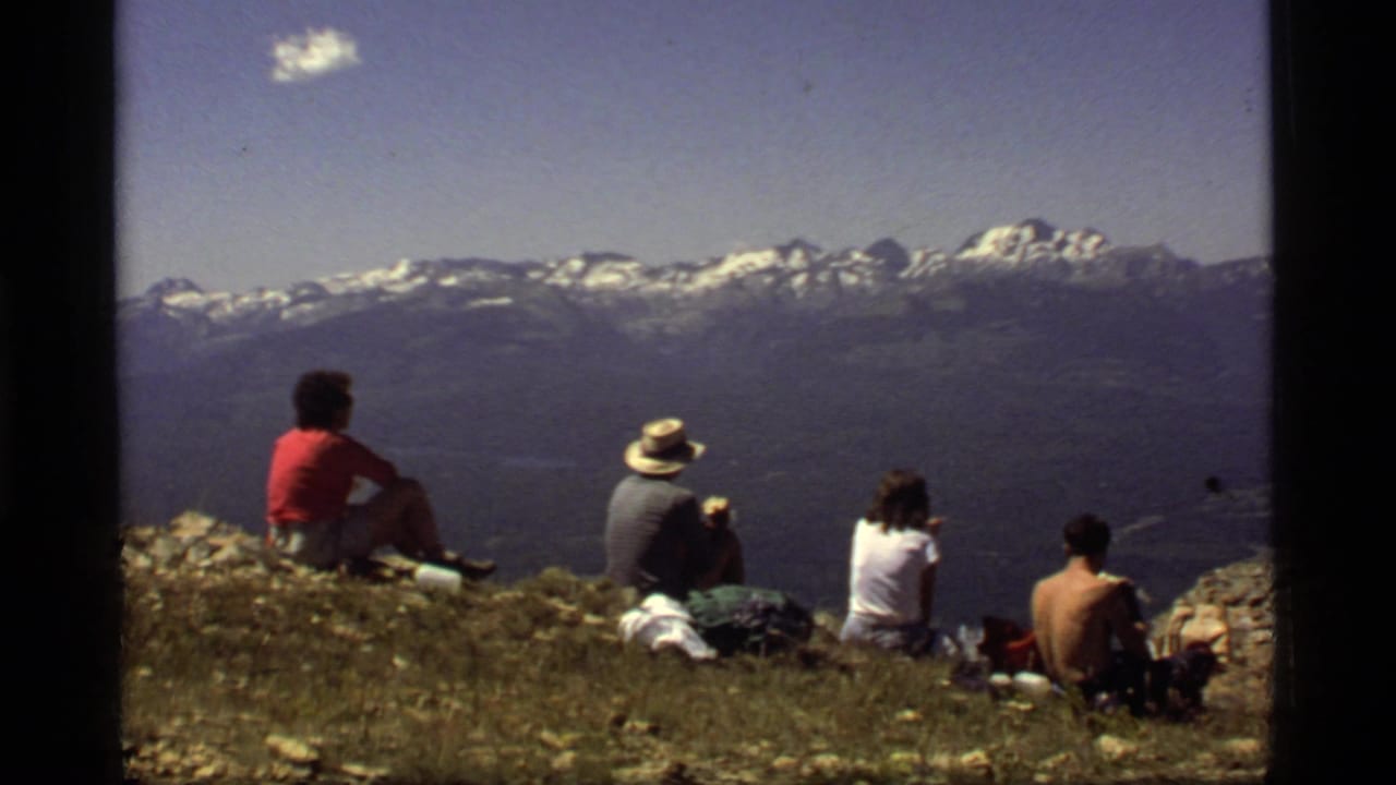 Seated hikers look out from a ridge toward a distant mountain range under blue sky.