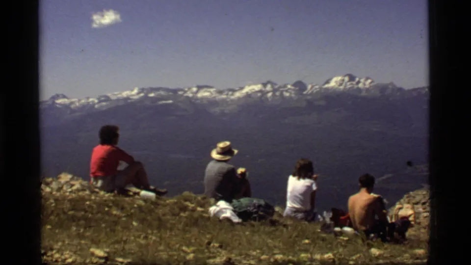 A group of hikers in the 1977 Montana home movie sit with backs to camera, overlooking a broad range of snow-streaked mountains.